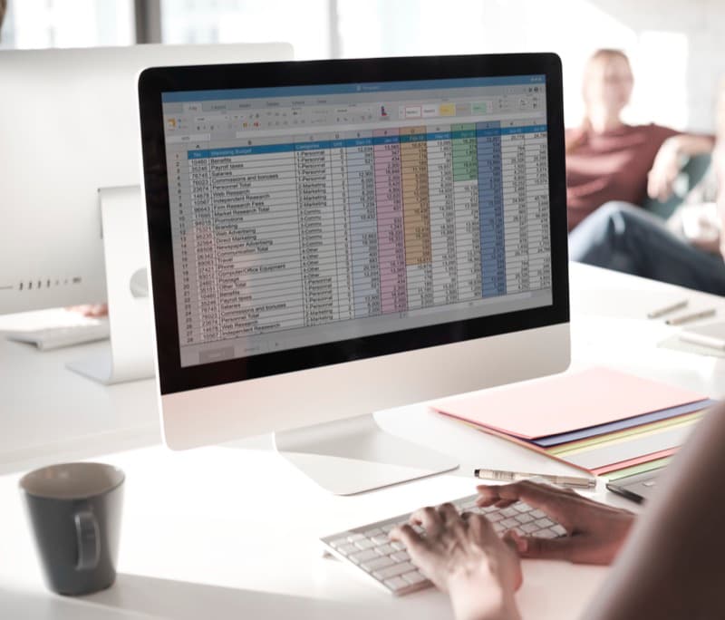 Person working on a desktop computer analyzing a marketing budget spreadsheet with various categories and amounts, in a bright office environment with colleagues in the background.