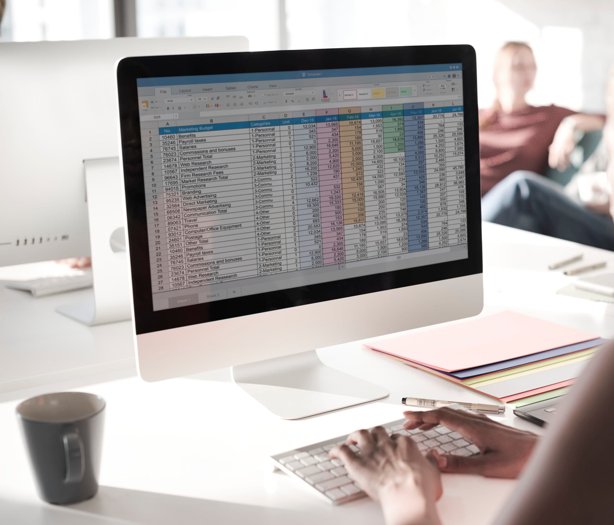 Person working on a desktop computer analyzing a marketing budget spreadsheet with various categories and amounts, in a bright office environment with colleagues in the background.